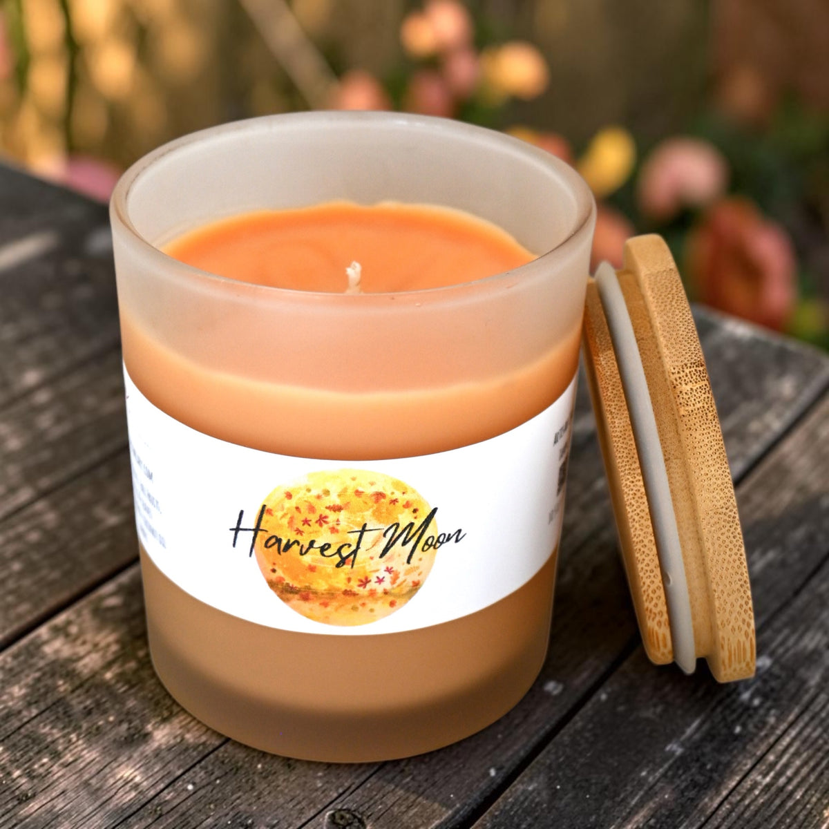 A frosted glass candle with orange wax sits on a dark wooden surface, with a bamboo lid leaning against it. The label reads 'Harvest Moon' and shows a warm-toned circular graphic.