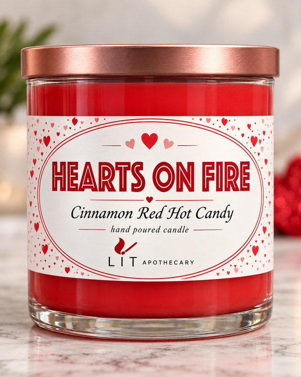 A glass jar candle with red wax and a metallic rose gold lid, featuring a white label with red hearts and the text 'HEARTS ON FIRE' and 'Cinnamon Red Hot Candy' by LIT APOTHECARY.
