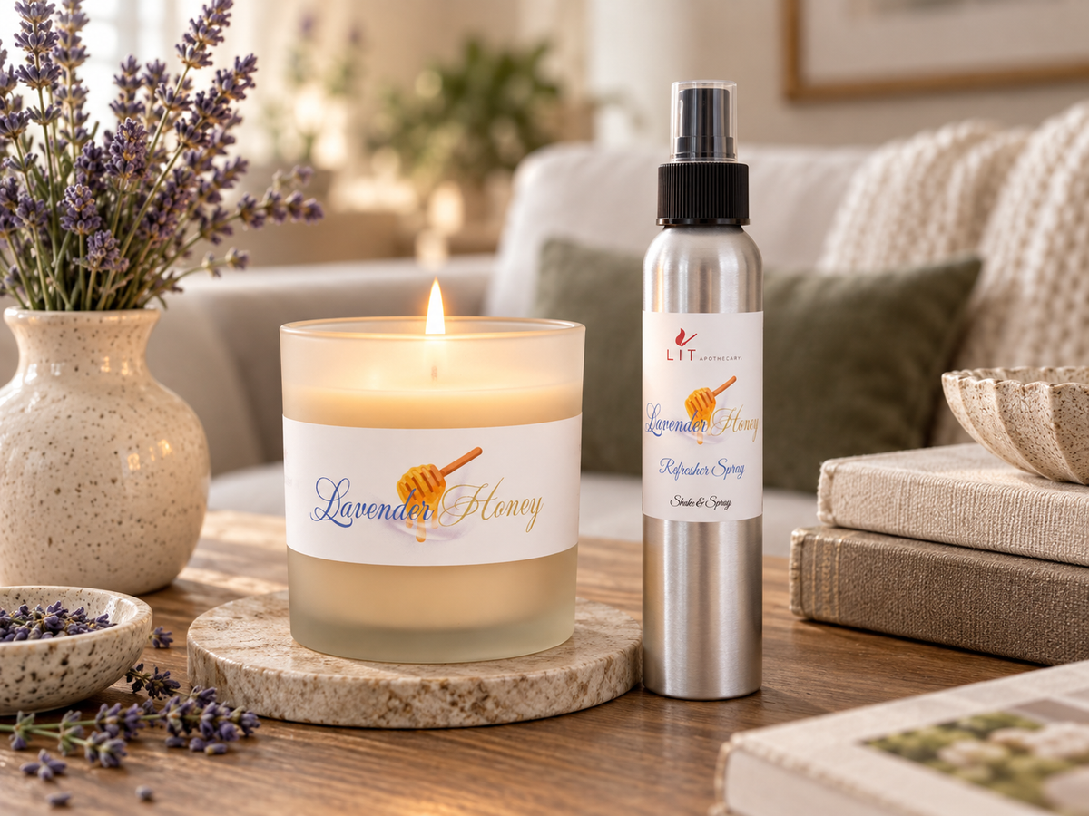 A lit lavender honey candle and a silver spray bottle labeled Lavender Honey Room Spray are displayed on a wooden table in a cozy living room setting with books, a vase of lavender, and a sofa in the background.