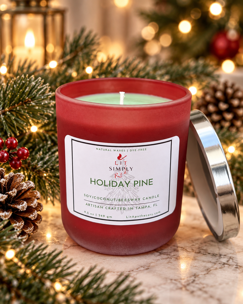 Holiday Pine candle in red frosted glass surrounded by evergreen branches, pinecones, and warm Christmas lights.