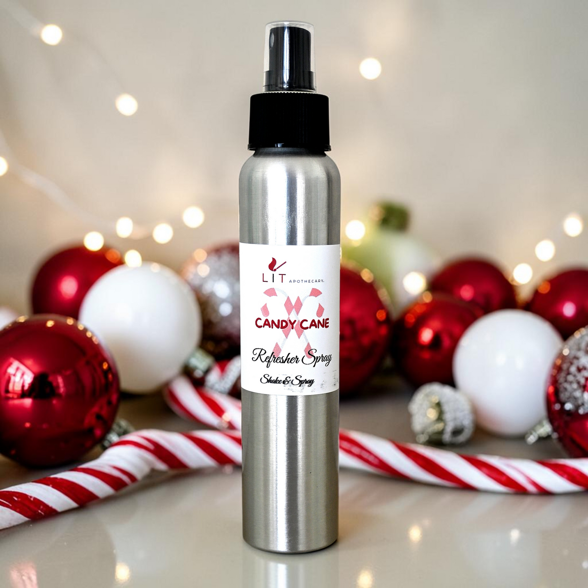 A silver aluminum spray bottle of LIT The Collection Candy Cane Refresher Spray stands on a glossy white surface, surrounded by festive red and white Christmas ornaments, candy canes, and warm string lights in the background.