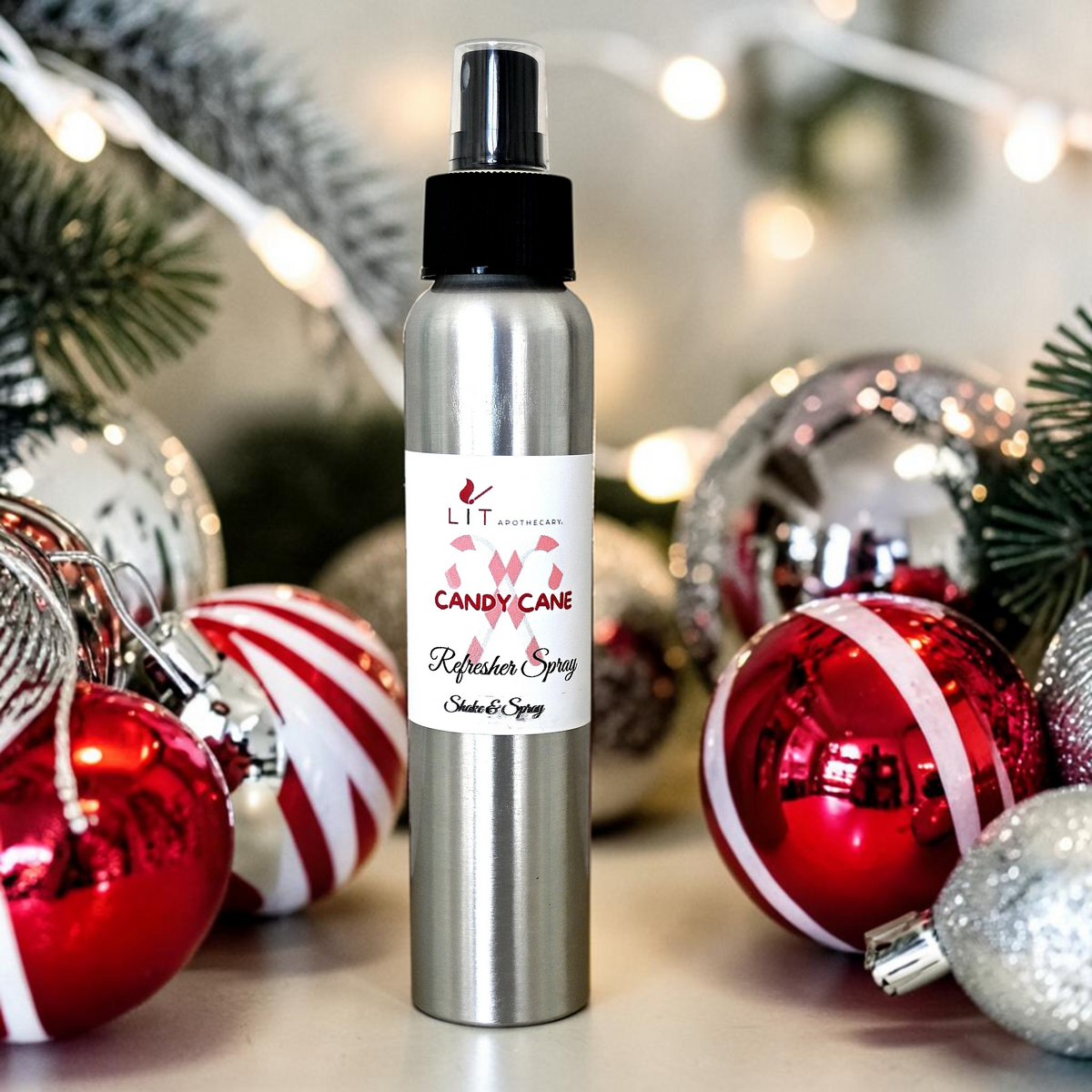 Lit Apothecary Candy Cane refresher spray surrounded by red, white and silver Christmas ornaments and warm holiday lights.