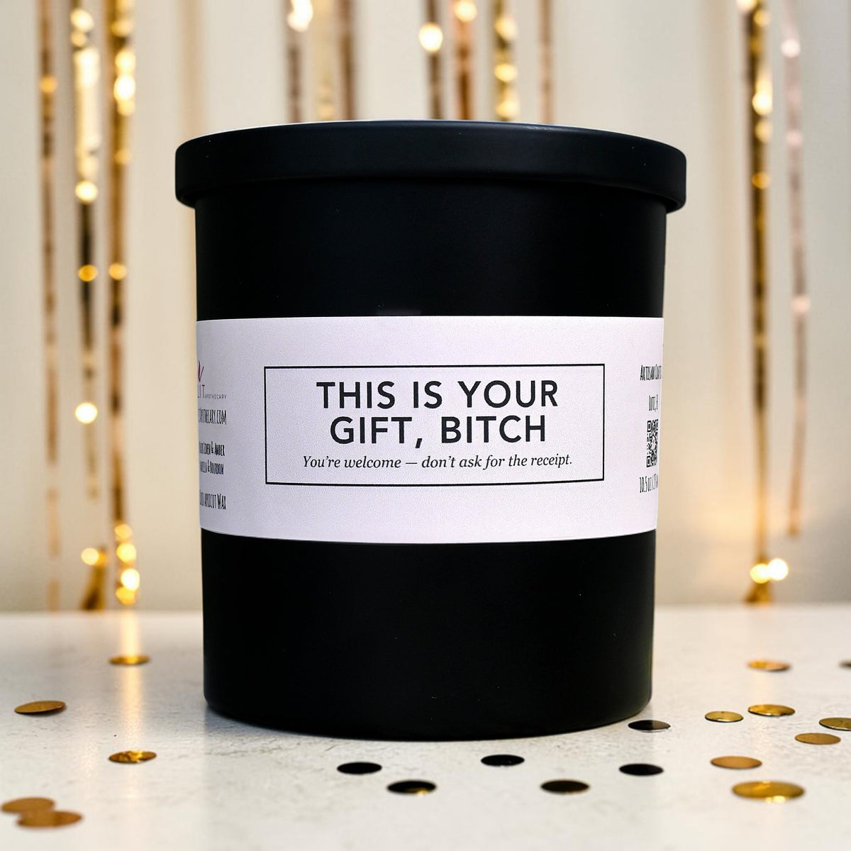 This Is Your Gift Bitch funny candle with black metal lid and cream label, front product photo in a gold party scene.