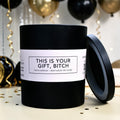 A matte black Lit Apothecary candle with its lid leaning against it, featuring a cream label that reads 'THIS IS YOUR GIFT, BITCH' with smaller text underneath. The box is photographed on a light surface with confetti, balloons, and streamers in the background.