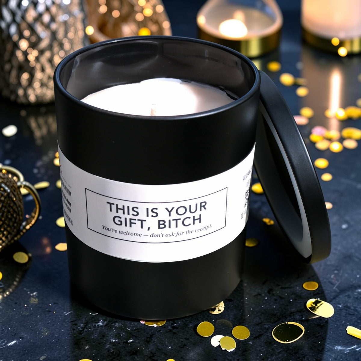 A black glass jar candle with its lid leaning against it, filled with cream wax and featuring a cream label that reads, 'THIS IS YOUR GIFT, BITCH.' The candle is styled on a dark reflective surface with scattered gold confetti and warm blurred candles in the background.