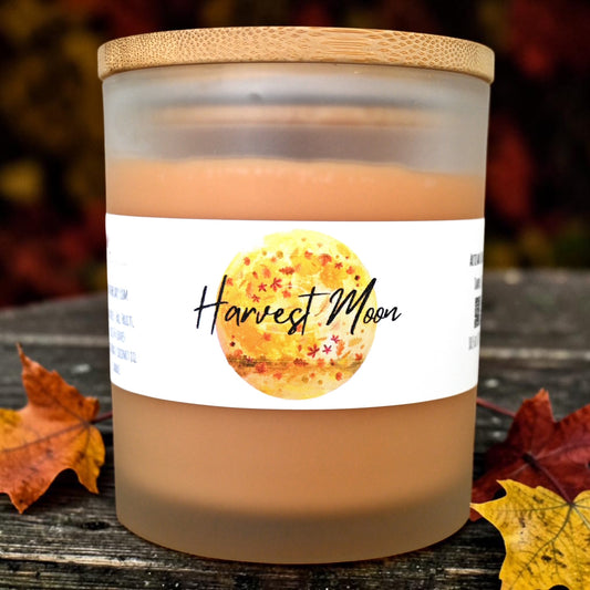 A frosted glass jar candle with a bamboo lid sits on a rustic wooden surface surrounded by autumn leaves. The candle wax is a warm peachy-brown color, and the label reads 'Harvest Moon' with a yellow-gold moon graphic.