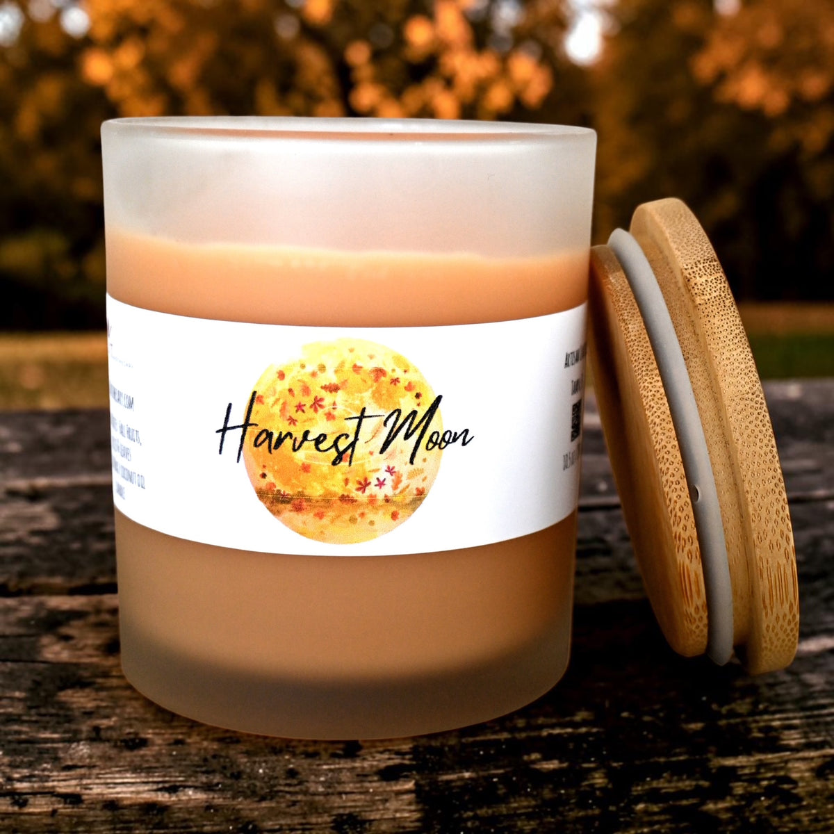 A frosted glass candle jar with a bamboo lid leaning against it on a rustic wooden surface, with a blurred outdoor background. The candle has a white label featuring a yellow-gold moon graphic and the text 'Harvest Moon'.
