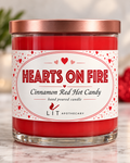 A glass jar candle with red wax and a metallic rose gold lid, featuring a white label with red hearts and the text 'HEARTS ON FIRE' and 'Cinnamon Red Hot Candy' by LIT APOTHECARY.