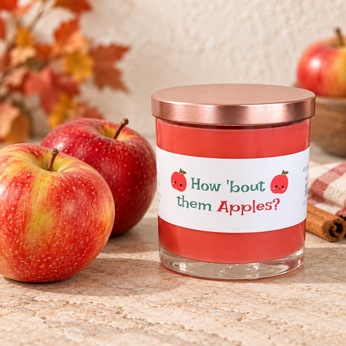 How ’Bout Them Apples scented candle with red wax, rose-gold lid, and fresh apples in a warm autumn setting.