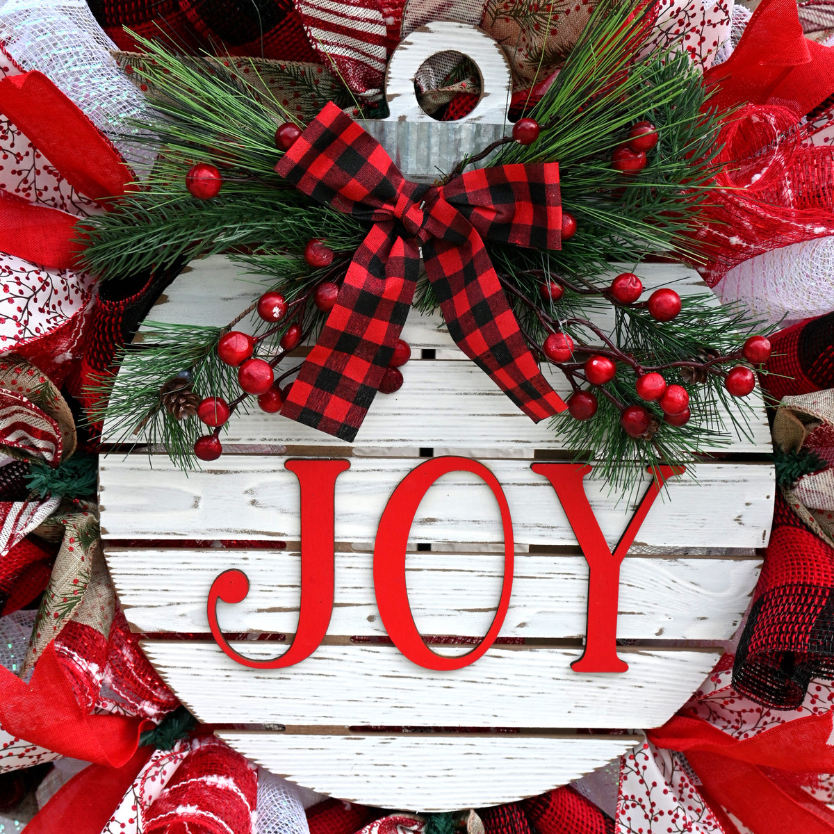 Close-up of white JOY ornament centerpiece on a Christmas wreath with greenery and festive holiday accents.