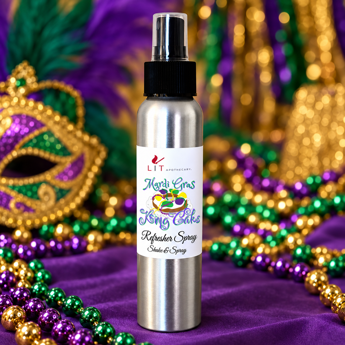 A silver metal spray bottle labeled 'LIT The Light Company Mardi Gras King Cake Refreshers Spray' placed on a purple fabric surface with Mardi Gras beads and festive masks in the background.