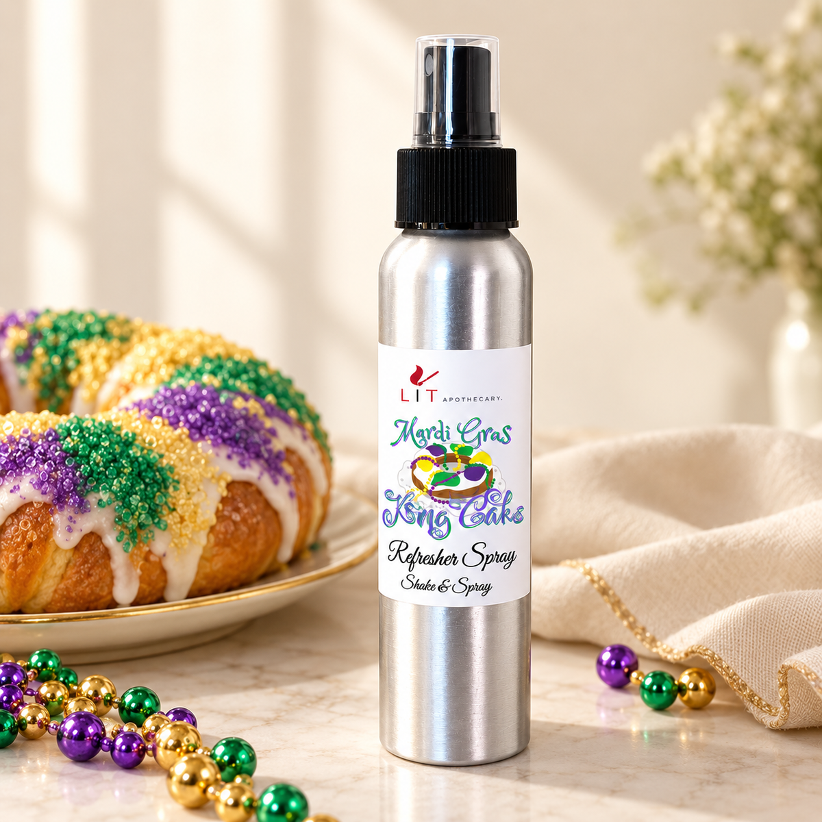 A silver metal spray bottle labeled 'LIT Apothecary Mardi Gras King Cake Refresher Spray' stands on a marble surface in a styled setting with a decorated King Cake, purple/green/gold beads, and a cream cloth in the background.