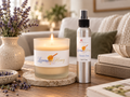 A lit lavender honey candle and a silver spray bottle labeled Lavender Honey Room Spray are displayed on a wooden table in a cozy living room setting with books, a vase of lavender, and a sofa in the background.