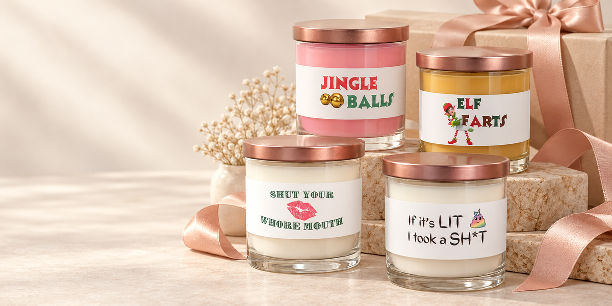 Four Lit Apothecary funny candles displayed on ivory stone risers with blush ribbon and a wrapped gift, including Jingle Balls, Elf Farts, and two everyday humor candles.
