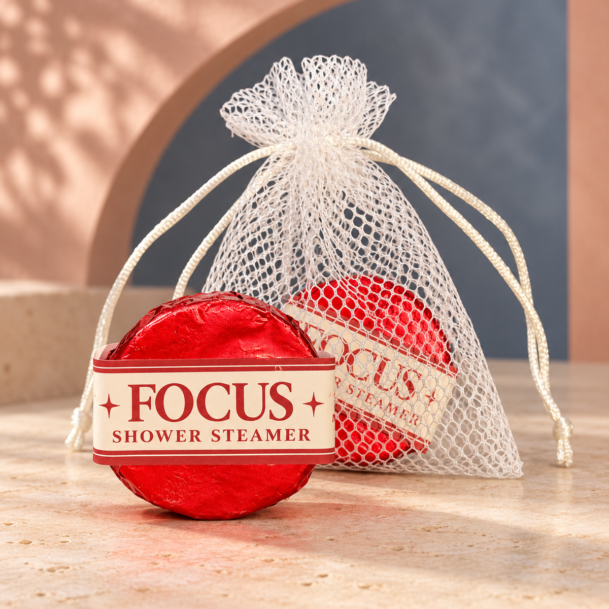 LIT Apothecary FOCUS shower steamer wrapped in red foil beside a white mesh pouch holding another steamer, displayed against a soft rose and slate-blue spa background.