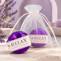 LIT Apothecary RELAX shower steamer wrapped in purple foil beside a white mesh pouch holding another steamer, displayed on pale stone against a soft lavender spa background.