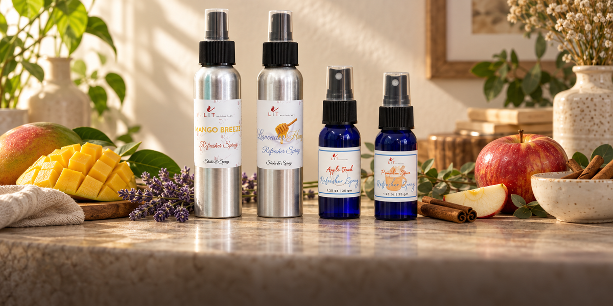 Four fragrance oil spray bottles of varying sizes arranged on a marble countertop with fresh ingredients like mango, apple, cinnamon, and lavender. The bottles are silver and cobalt blue with white labels and black spray nozzles.