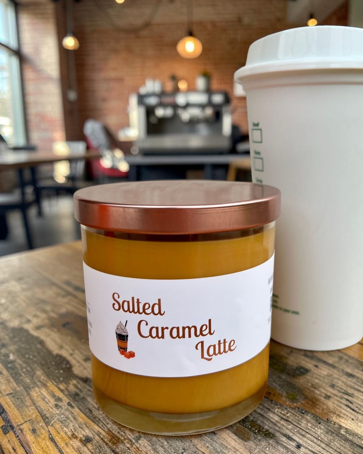 A jar candle labeled 'Salted Caramel Latte' with amber-brown wax and a metallic rose-gold lid, sitting on a wooden table in a cafe setting with a takeaway coffee cup in the background.