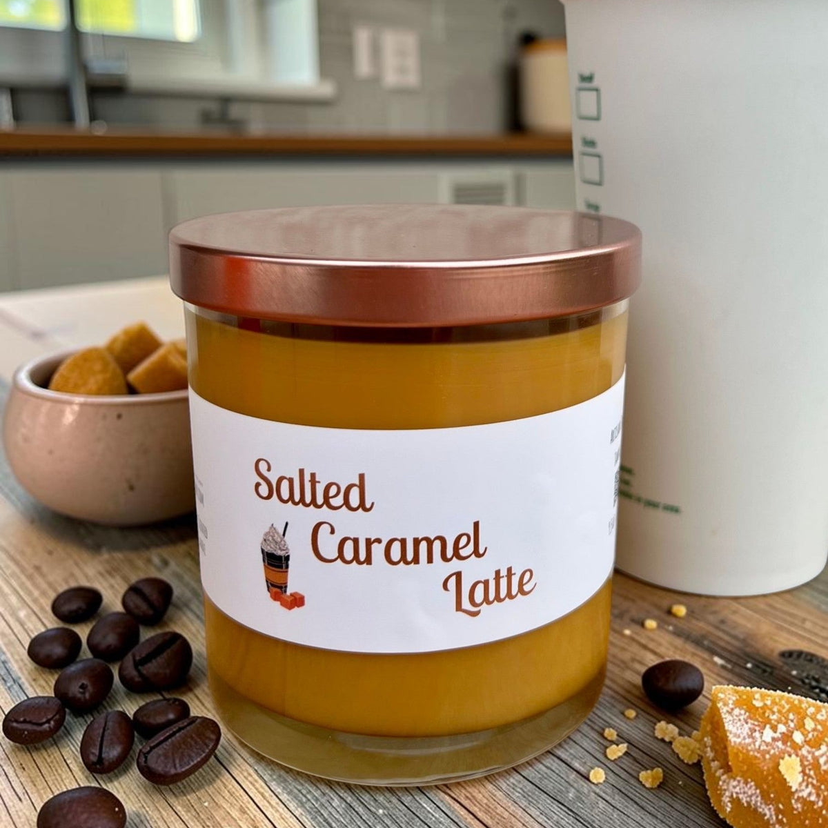 A candle in a glass jar with a white label reading 'Salted Caramel Latte' and a metallic copper lid, styled on a wooden surface with coffee beans, a bowl of sugar, and a white container in a kitchen setting.