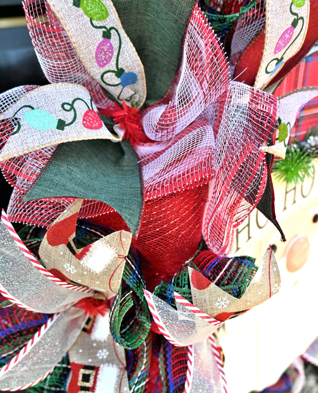 Close-up of layered Christmas wreath ribbons with holiday lights, plaid, stockings, glitter accents, and classic red and green colors.