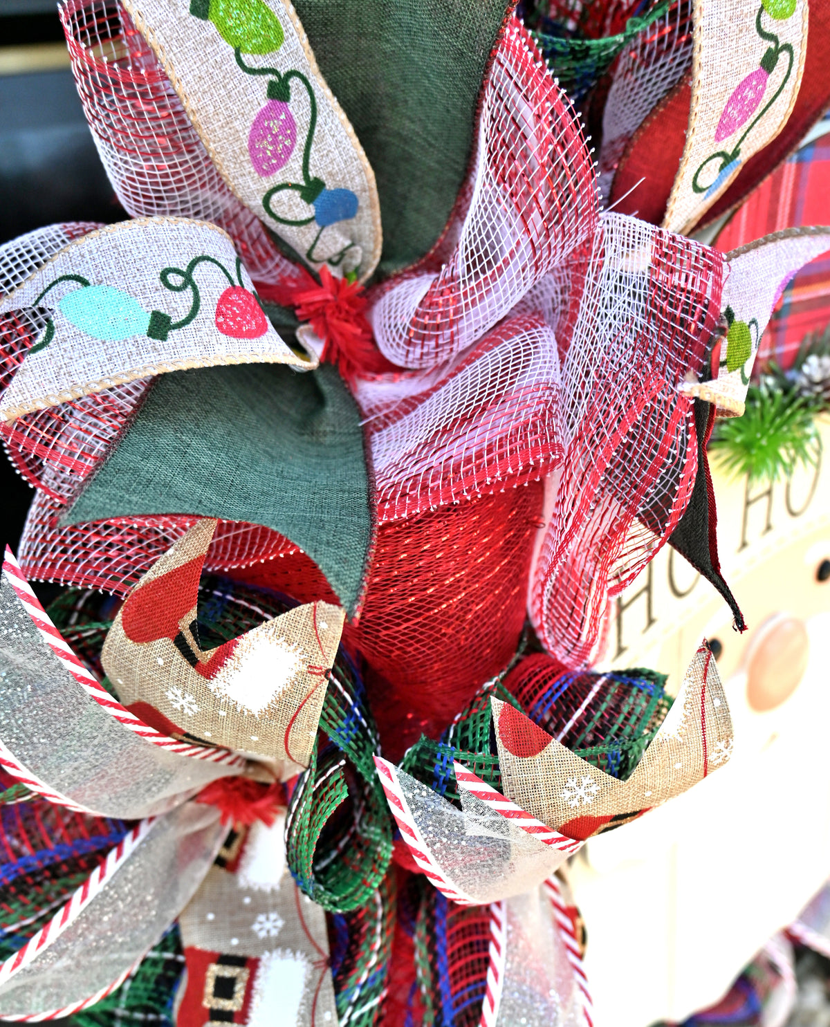 Close-up of layered Christmas wreath ribbons with holiday lights, plaid, stockings, glitter accents, and classic red and green colors.