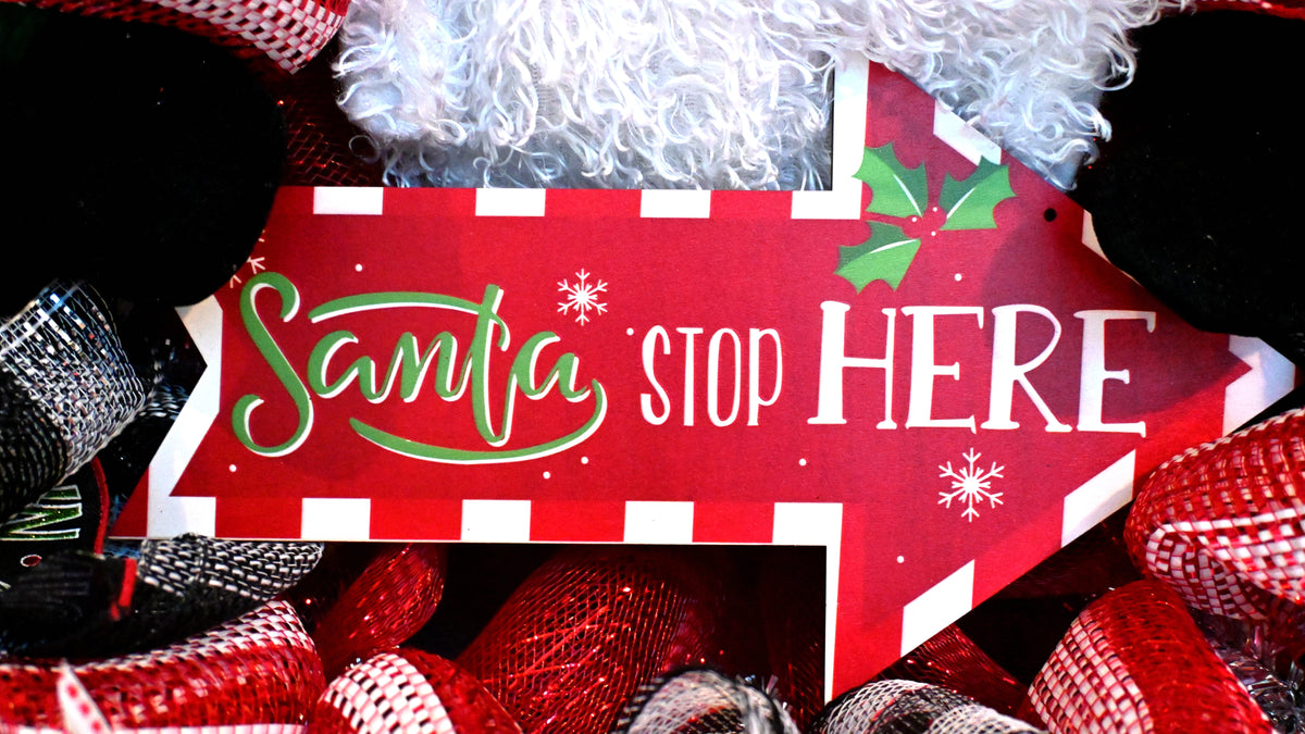 Close-up of Santa Stop Here sign on a Christmas wreath with festive red, white and black holiday accents.