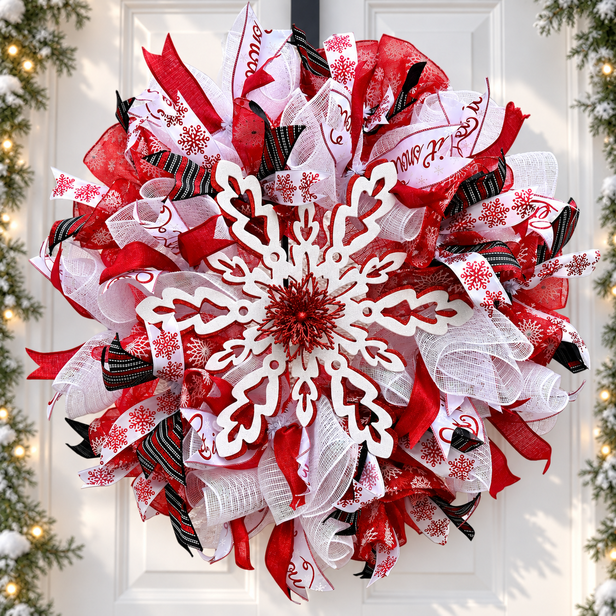 A festive red, white, and black mesh deco wreath with a large white snowflake cutout in the center, hanging on a white front door. The wreath features red, white, and black ribbons with holiday patterns, and is framed by blurred evergreen branches with warm lights on the sides.