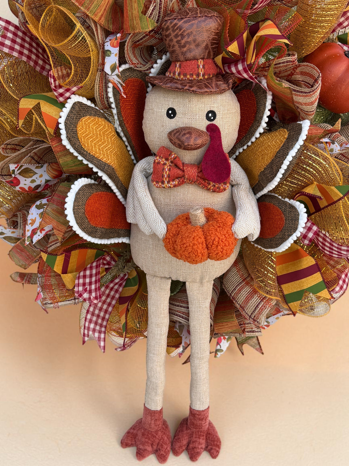 Close-up of the plush turkey wearing a brown top hat and plaid bow tie, holding an orange pumpkin on the Thanksgiving wreath