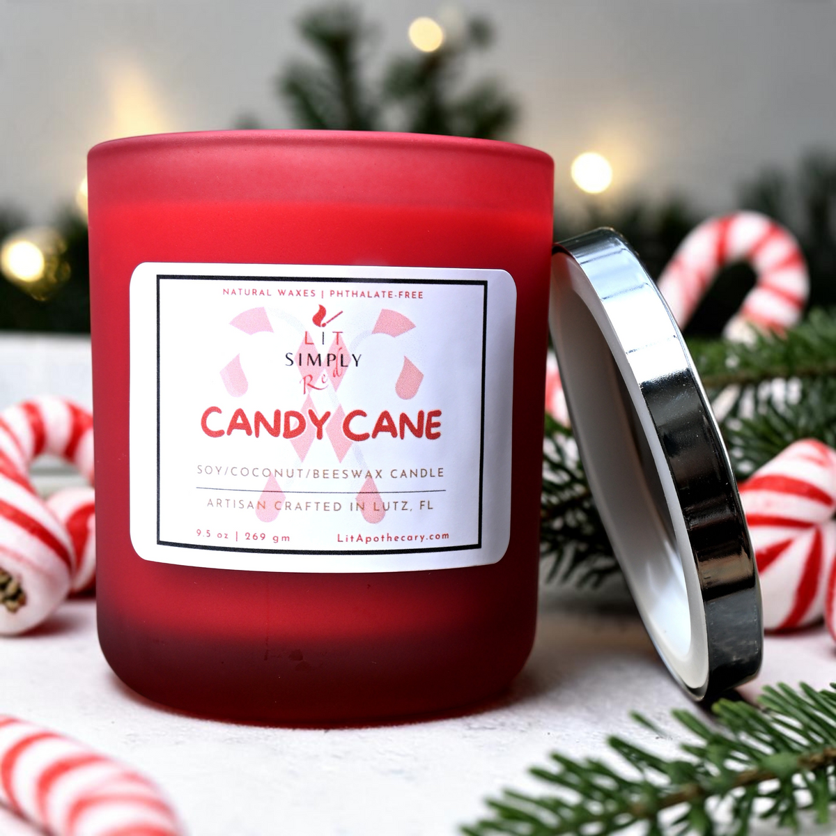 Open Candy Cane candle in a red frosted glass jar with its silver lid beside it, surrounded by peppermint candy and evergreen accents.