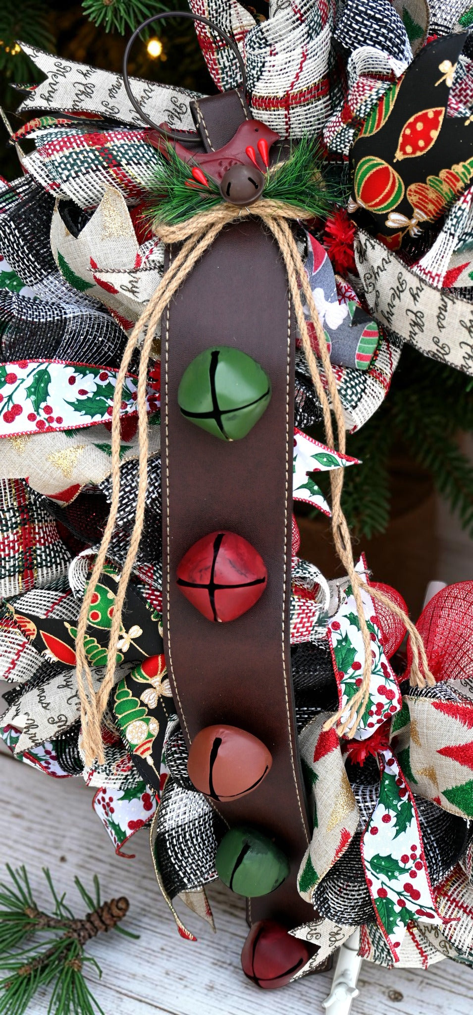 Close-up of the wreath's jingle bells on a stitched brown leather strap with twine, greenery, and a red bird