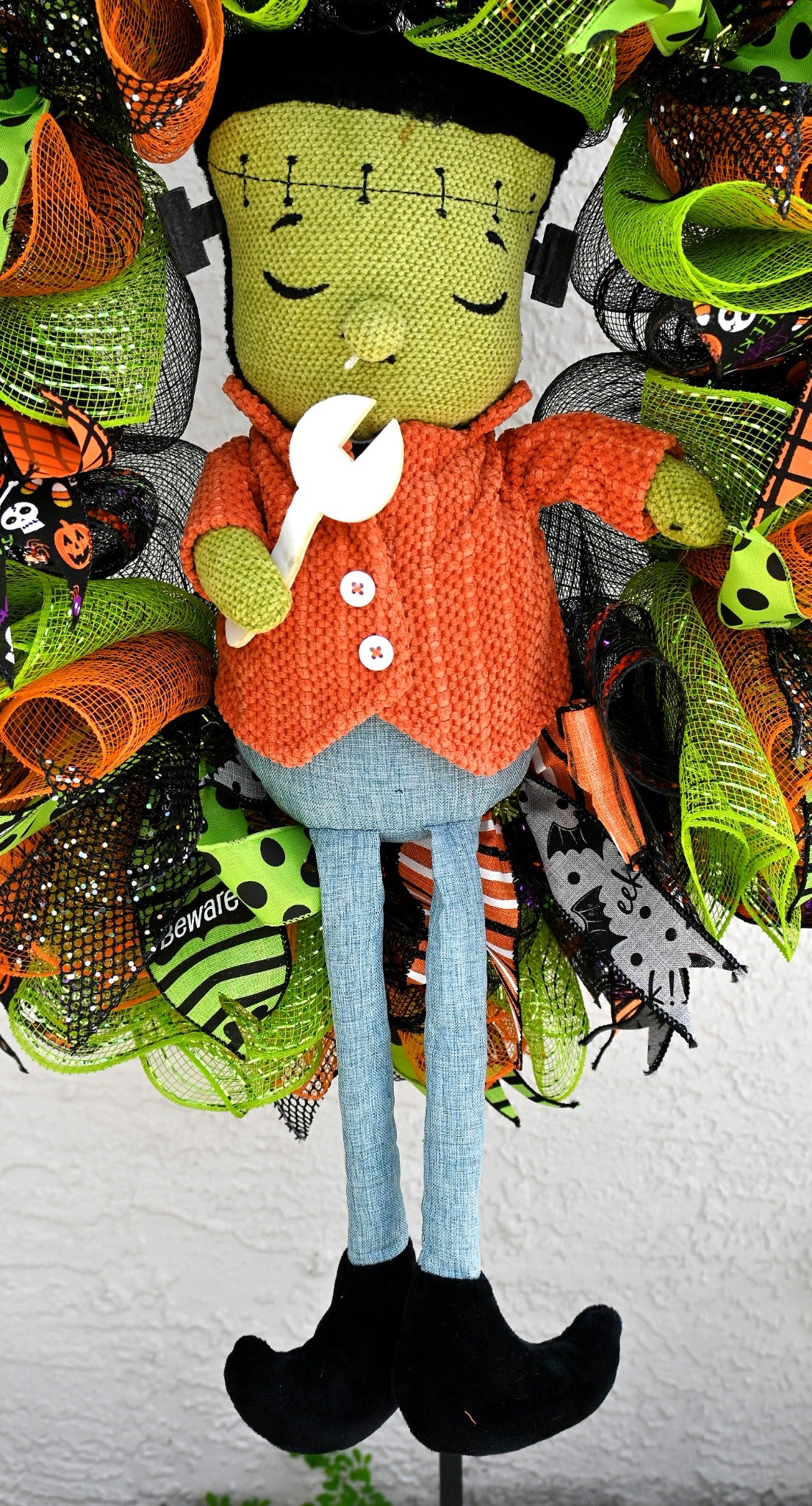 Close-up of crocheted Frankenstein figure with orange jacket, wrench and long blue legs on Halloween wreath