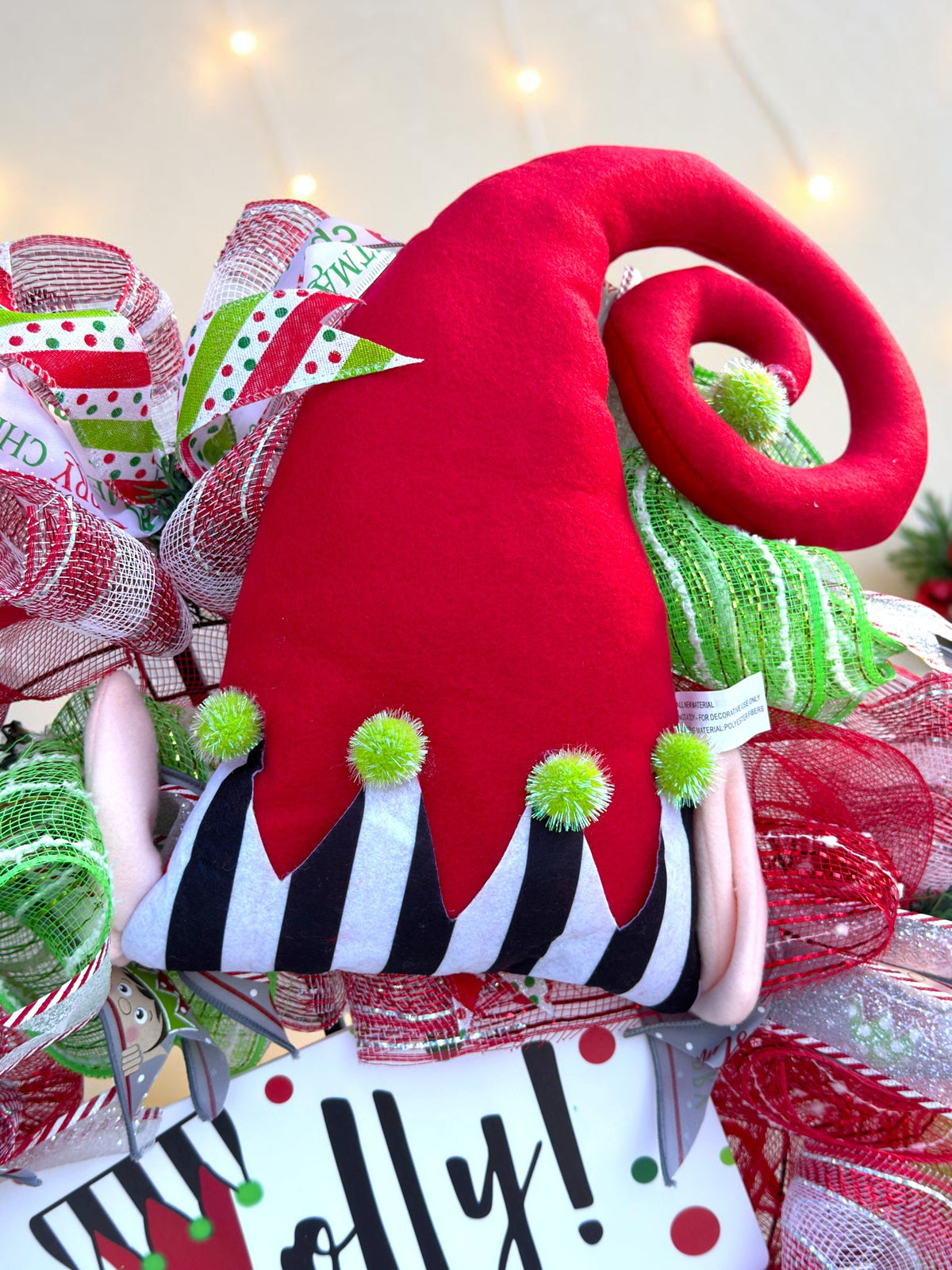 Close-up of red curled elf hat with striped brim and green pom-poms on Christmas wreath.